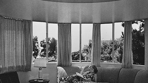 A woman sitting on the couch reading in front of a window.